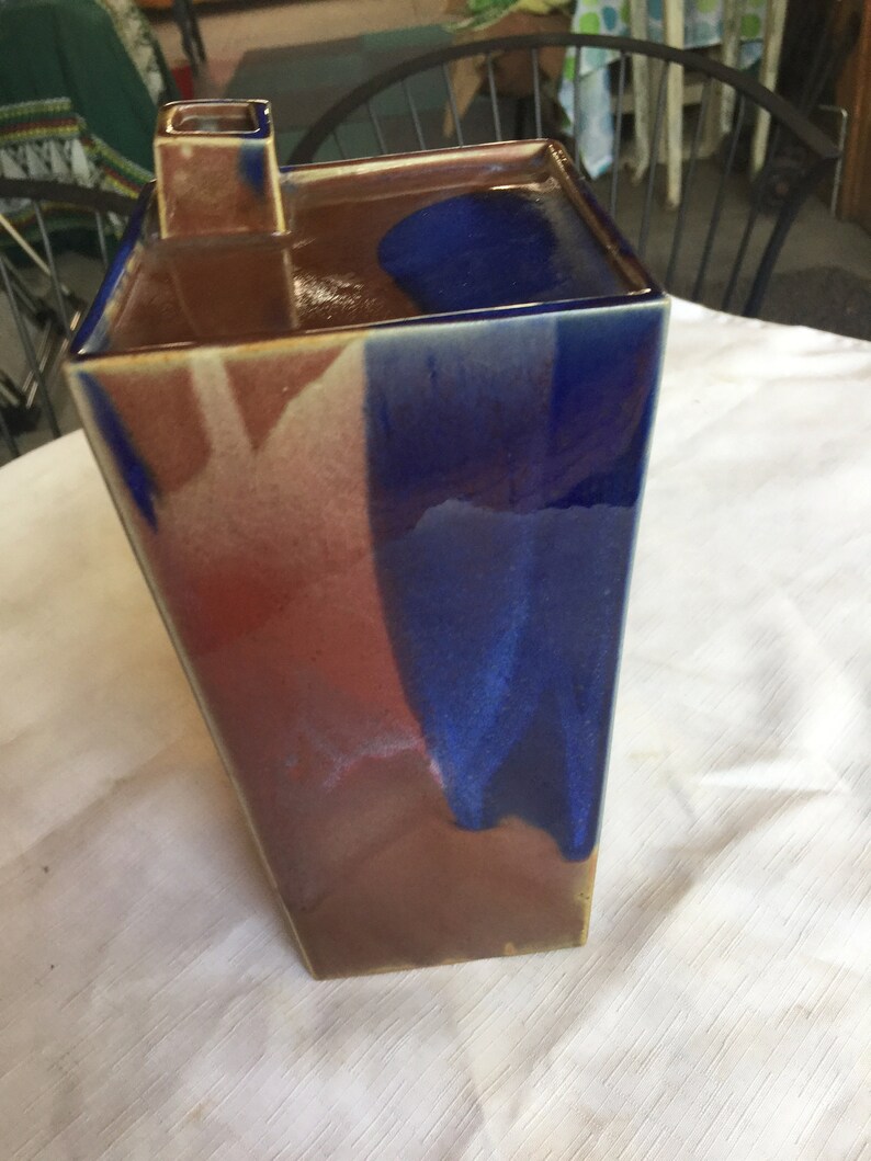 Op de afbeelding: Een hoge, rechthoekige keramische vaas met een vierkante opening aan de bovenkant. De vaas is geglazuurd in tinten bruin, rood en blauw, wat een marmerachtig effect cre&euml;ert.