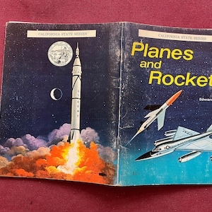 May include: A vintage book titled "Planes and Rockets" from the California State Series, featuring a rocket launching into space and a jet plane flying in the sky. The book is by Edward Victor.