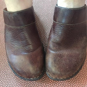 May include: A pair of brown leather clogs with a white stitch detail around the top edge. The clogs have a worn look and are made of a textured leather.