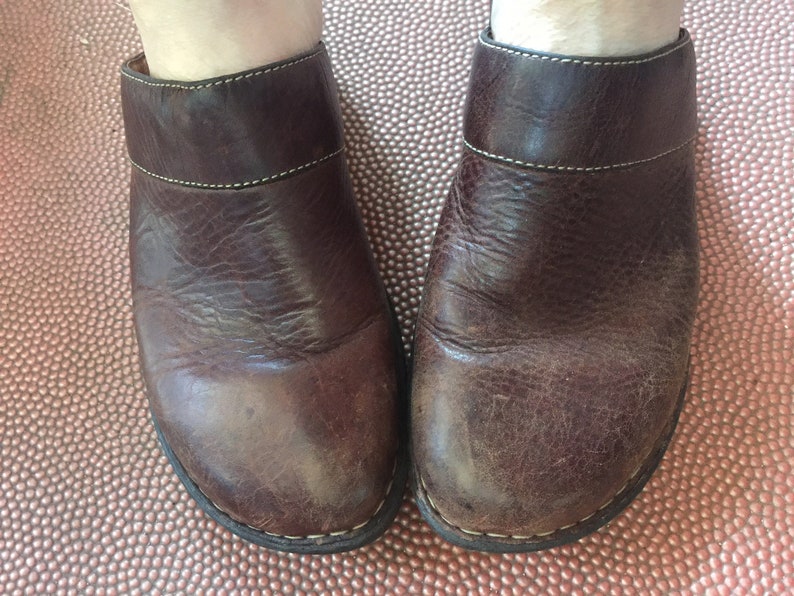 May include: A pair of brown leather clogs with a white stitch detail around the top edge. The clogs have a worn look and are made of a textured leather.