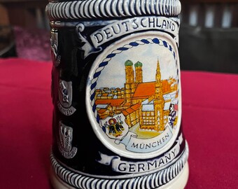 May include: A white ceramic beer stein with a blue and white design. The stein features a depiction of the city of Munich, Germany, with the text "DEUTSCHLAND" and "GERMANY" on the front.