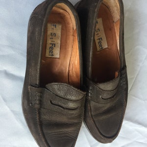 Może przedstawiać: Para brązowych sk&oacute;rzanych mokasyn&oacute;w z etykietą "Free S Feet" wewnątrz każdego buta.