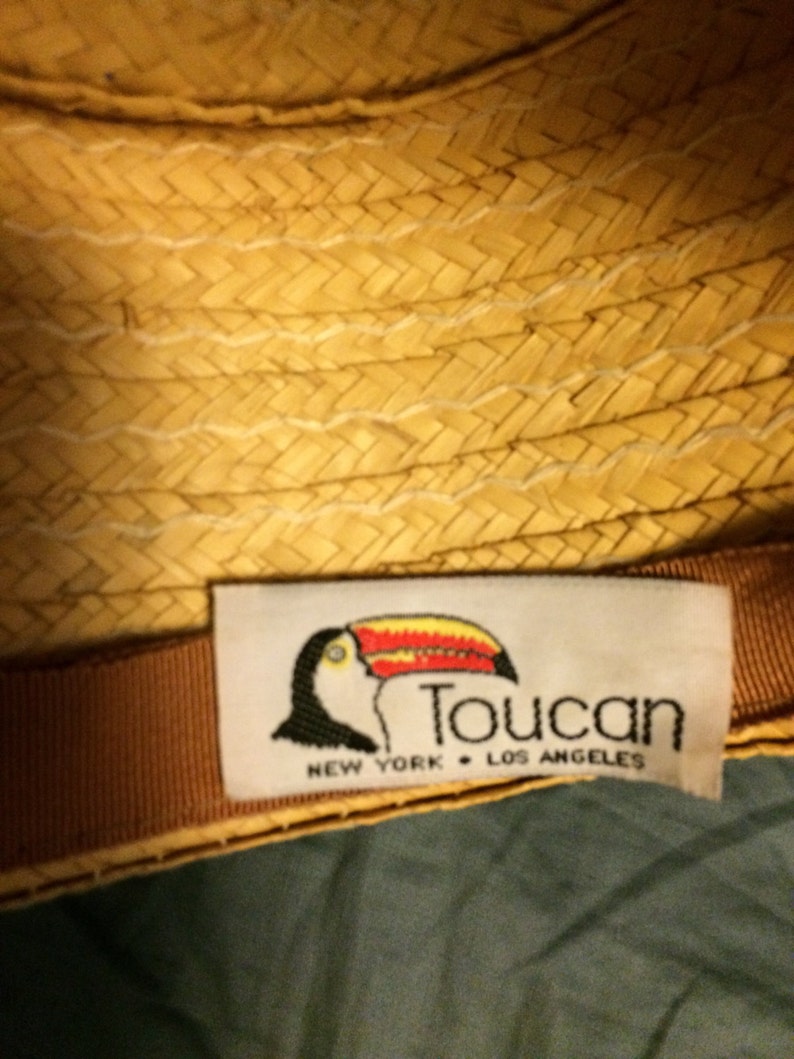 Pu&ograve; includere: Un cappello di paglia intrecciato marrone e beige con un nastro marrone e un'etichetta bianca con un'illustrazione di tucano e il testo "Toucan New York Los Angeles".