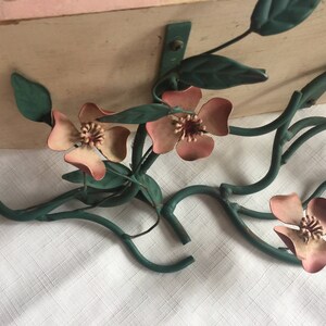 May include: A metal wall shelf with a floral design. The shelf is painted in a light pink color and features a green metal floral design with pink flowers. The shelf is attached to a white wall.