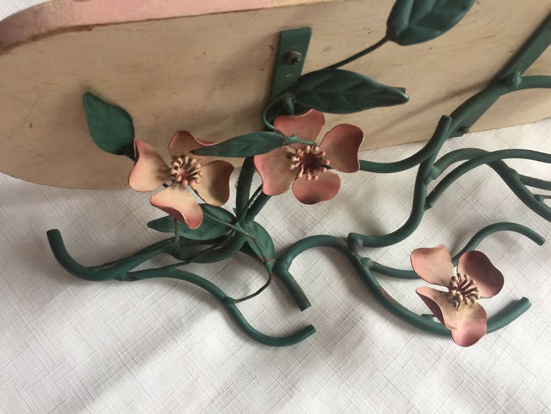 May include: A metal wall shelf with a floral design. The shelf is painted in a light pink color and features a green metal floral design with pink flowers. The shelf is attached to a white wall.