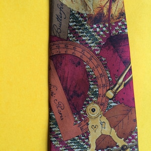 May include: A brown, red, and green patterned silk tie with a compass and protractor design. The tie features the text "Butterflie" and "Civid. Paris".