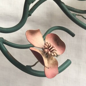 May include: A close-up of a pink metal flower with a green metal stem. The flower has five petals and a center with a raised design. The stem is twisted and has a rustic look.