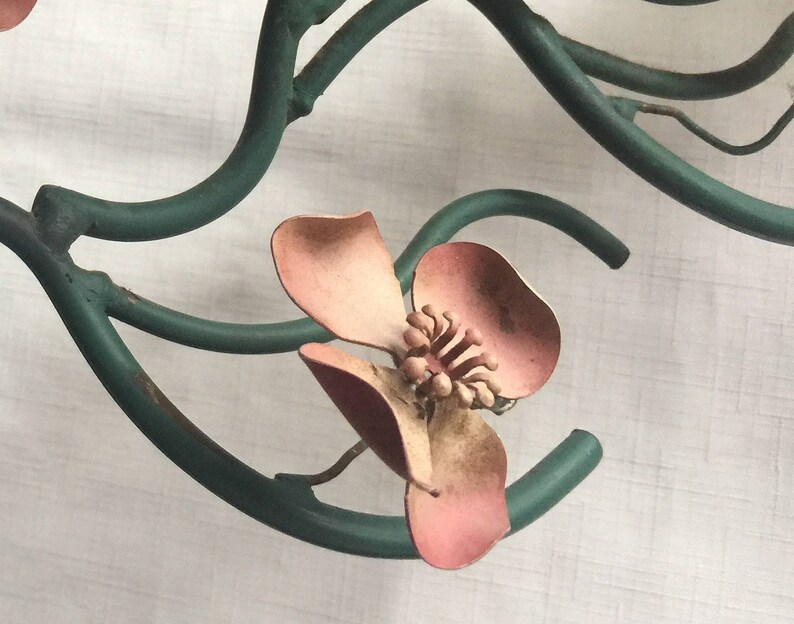 May include: A close-up of a pink metal flower with a green metal stem. The flower has five petals and a center with a raised design. The stem is twisted and has a rustic look.