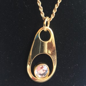 May include: A gold pendant necklace with a large, clear crystal in the center. The pendant is shaped like a teardrop with a rounded top and a pointed bottom. The chain is a delicate, gold chain.