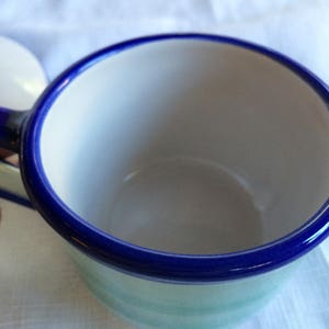 May include: A white ceramic teacup with a blue rim and a blue handle. The cup is empty and has a light blue and white pattern on the outside.