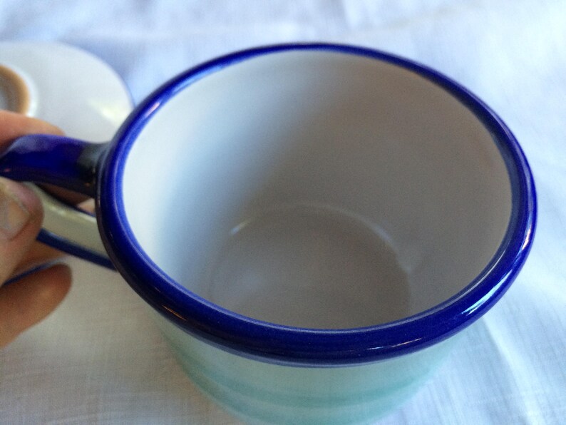 May include: A white ceramic teacup with a blue rim and a blue handle. The cup is empty and has a light blue and white pattern on the outside.