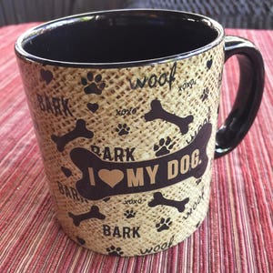 Könnte beinhalten: Eine schwarze Keramik-Kaffeetasse mit einem braunen, beige und schwarzen Design. Die Tasse hat ein Knochen-, Pfotenabdruck- und Herzmuster mit den Worten "woof", "bark" und "I love my dog".