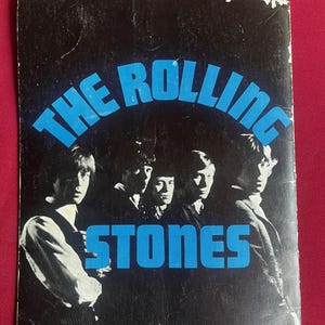 Puede incluir: Libro de recuerdos vintage de los Rolling Stones en negro y azul. El nombre de la banda está en letras azules grandes, con una foto de los miembros de la banda debajo. El libro está sobre un fondo rojo.