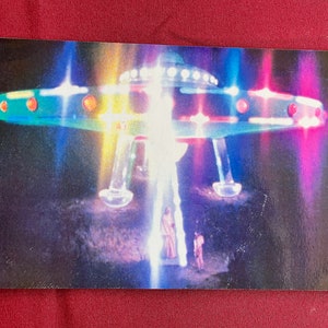 Könnte beinhalten: Eine Vintage-Postkarte mit einem farbenfrohen Bild einer fliegenden Untertasse mit leuchtenden Lichtern, die auf einer dunklen, felsigen Oberfläche landet. Zwei Figuren stehen im Vordergrund, beleuchtet von den Strahlen der Untertasse.