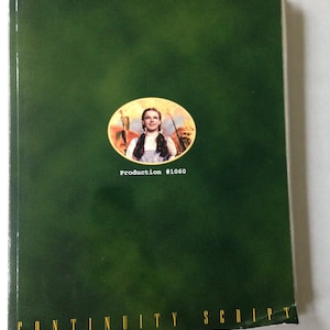Puede incluir: Un libro verde con el título "El mago de Oz" y el texto "Continuity Script" en la portada. Una imagen circular de Judy Garland como Dorothy de la película está en el centro de la portada. El texto "Production #1060" está debajo de la imagen.