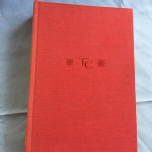 Könnte beinhalten: Ein rotes Hardcover-Buch mit strukturierter Oberfläche. Auf dem Einband sind die Initialen "TC" und zwei dekorative Symbole abgebildet. Das geschlossene Buch ist in gutem Zustand. Die Farbe ist ein leuchtendes Rot.