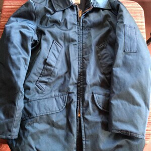May include: A dark blue jacket with a zipper closure and two pockets on the front. The jacket has a patch on the right sleeve.