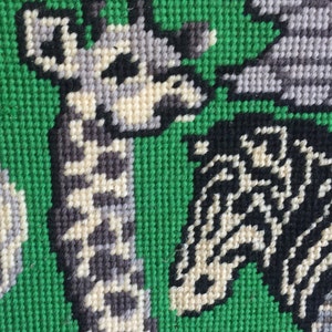 May include: A framed needlepoint picture of a giraffe, zebra, and elephant on a green background. The animals are stitched in black, white, and gray yarn.