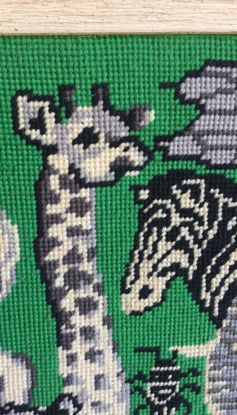 May include: A framed needlepoint picture of a giraffe, zebra, and elephant on a green background. The animals are stitched in black, white, and gray yarn.