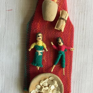 May include: A handmade wall hanging featuring two small yarn dolls, a small clay pot with a feather plume, a woven rug, and a bowl of white ceramic chips on a wooden base.