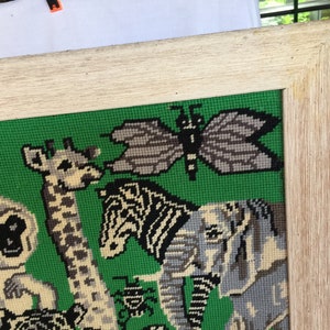 May include: A framed needlepoint picture with a green background featuring a giraffe, zebra, elephant, butterfly, and several beetles. The frame is a light wood color.