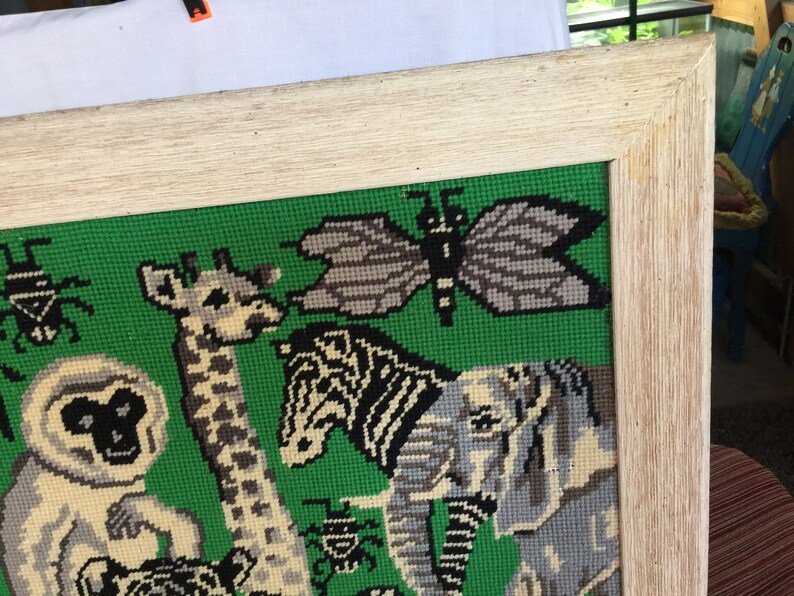 May include: A framed needlepoint picture with a green background featuring a giraffe, zebra, elephant, butterfly, and several beetles. The frame is a light wood color.