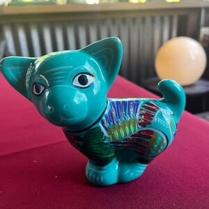 May include: A teal blue ceramic figurine of a cat with colorful painted details. The cat has large eyes and a whimsical expression.