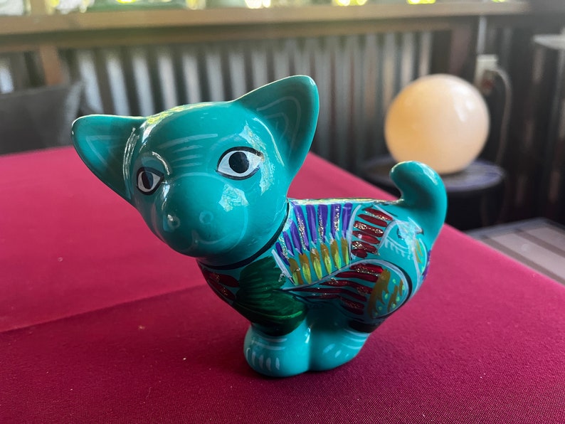 May include: A teal blue ceramic figurine of a cat with colorful painted details. The cat has large eyes and a whimsical expression.