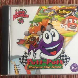 May include: A CD case for the computer game Putt-Putt Enters the Race. The case features a cartoon image of Putt-Putt, a purple car with a smiling face, driving on a race track. The game is part of the It's a Junior Adventure series for kids ages 3-8.