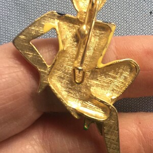 Puede incluir: Broche dorada con una figura estilizada de una mujer sentada con las piernas cruzadas. La broche tiene un dise&ntilde;o de esmalte verde y amarillo y un cierre de pin.