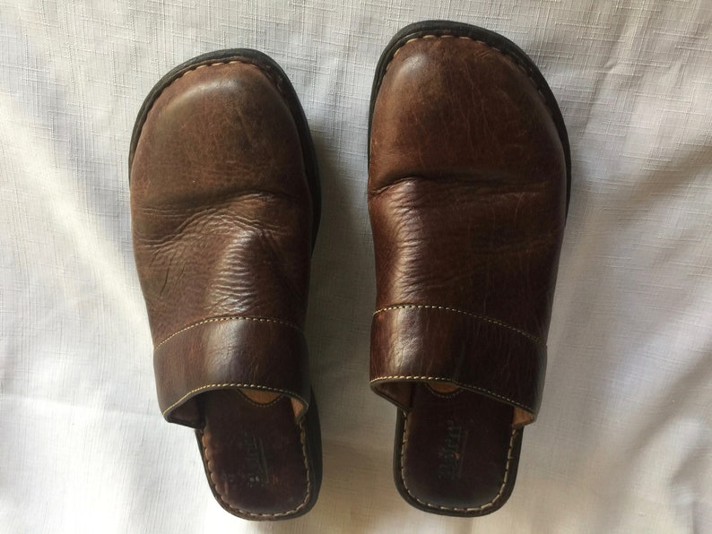 May include: A pair of brown leather slip-on shoes with a closed toe and a back strap. The shoes have a brown leather sole and a brown leather upper.