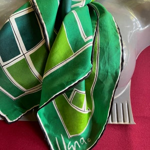 May include: A green and white silk scarf with a geometric pattern and black trim. The scarf has the word "Vana" written on it.