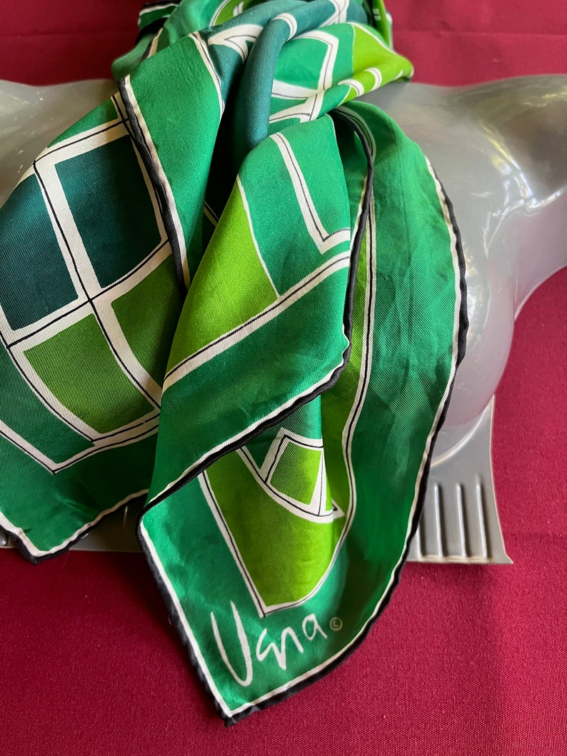 May include: A green and white silk scarf with a geometric pattern and black trim. The scarf has the word "Vana" written on it.