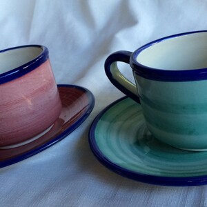 May include: Two ceramic teacups and saucers. The teacup on the left is pink with a blue rim. The teacup on the right is green with a blue rim. Both saucers have a blue rim and a swirl pattern.