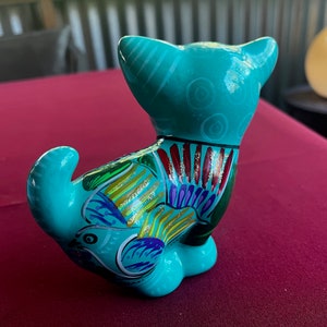 May include: A turquoise ceramic cat figurine with colorful painted details. The cat is sitting with its tail curled around its body. The figurine has a glossy finish.