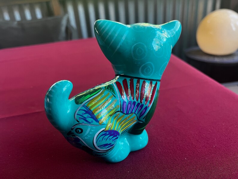 May include: A turquoise ceramic cat figurine with colorful painted details. The cat is sitting with its tail curled around its body. The figurine has a glossy finish.