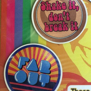 May include: Two round coasters with colourful designs. The first coaster is brown with a white and pink design that says "Shake it, don't break it". The second coaster is red, yellow, and blue with the words "Far Out" in blue.
