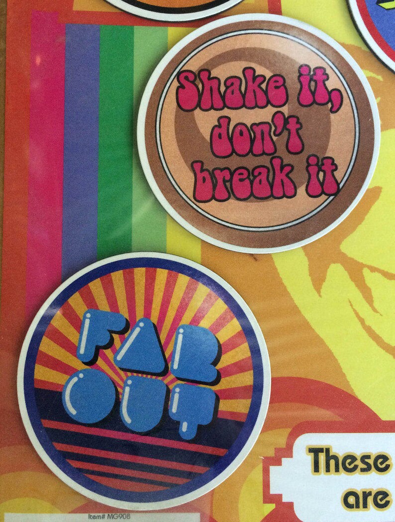May include: Two round coasters with colourful designs. The first coaster is brown with a white and pink design that says "Shake it, don't break it". The second coaster is red, yellow, and blue with the words "Far Out" in blue.