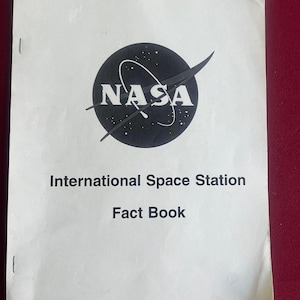 May include: A white booklet featuring the NASA logo, a black circle with white text and a curved line. The text "International Space Station Fact Book" is printed below the logo in black.