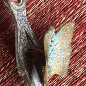 May include: An antique metal object with a butterfly wing design. The object has a weathered appearance with gold, blue, and silver tones. The intricate details and aged patina suggest it is a vintage decorative piece.