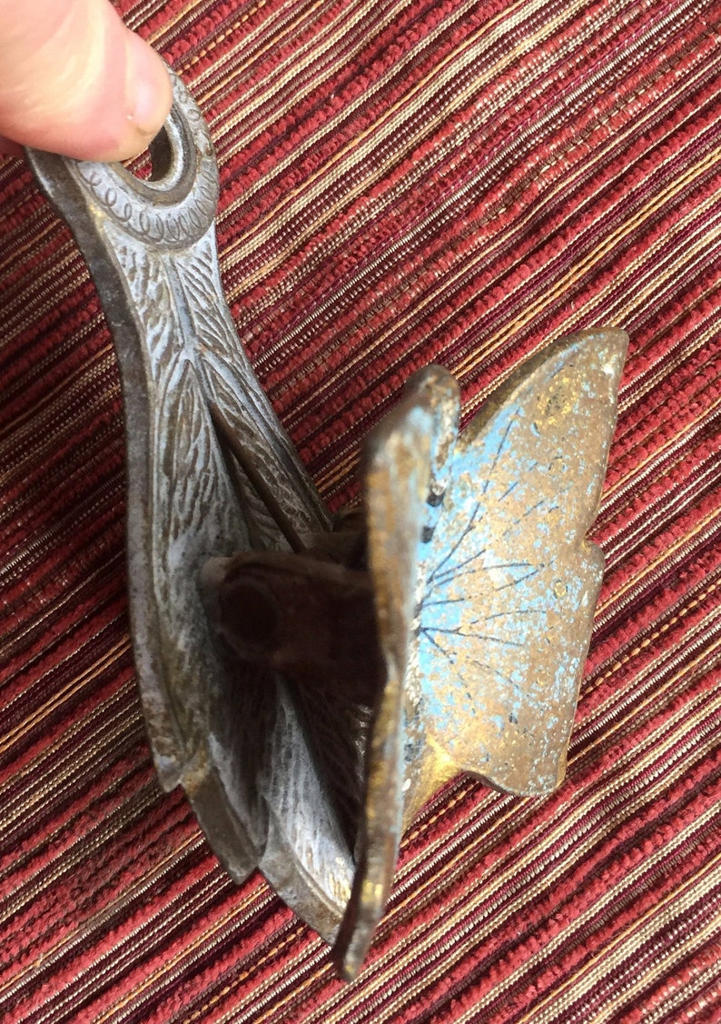 May include: An antique metal object with a butterfly wing design. The object has a weathered appearance with gold, blue, and silver tones. The intricate details and aged patina suggest it is a vintage decorative piece.
