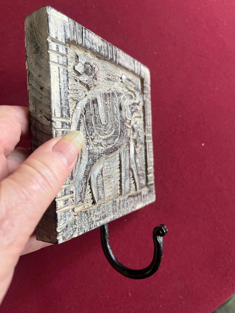 May include: A square, wooden wall hook with a carved elephant design. The hook is painted in shades of gray and white, with a black metal hook. The elephant carving is detailed and the hook is attached to the bottom of the square.