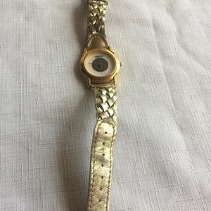 May include: A gold-toned wristwatch with a white face and a braided gold-toned band. The watch face has a circular design with a small circle in the center.