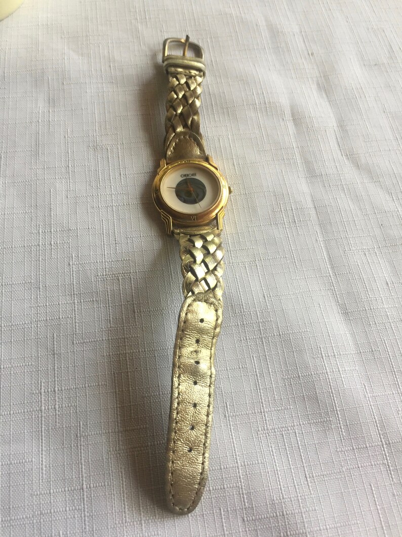 May include: A gold-toned wristwatch with a white face and a braided gold-toned band. The watch face has a circular design with a small circle in the center.