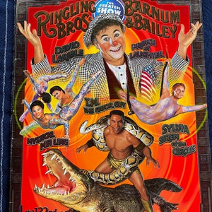 Peut inclure: Une affiche rouge et or pour le cirque Ringling Bros. et Barnum & Bailey. L'affiche présente un clown, un charmeur de serpents, un artiste mystique et un alligator. Le texte sur l'affiche indique "The Greatest Show" et "The 132nd Circus".