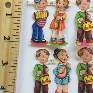 Peut inclure: Six poup&eacute;es en papier vintage repr&eacute;sentant des enfants jouant d'instruments de musique. Les poup&eacute;es sont habill&eacute;es de v&ecirc;tements color&eacute;s et tiennent des instruments tels qu'un tambour, un accord&eacute;on, un saxophone et un banjo. Les poup&eacute;es sont imprim&eacute;es sur du papier blanc et portent une &eacute;tiquette "Made in England".