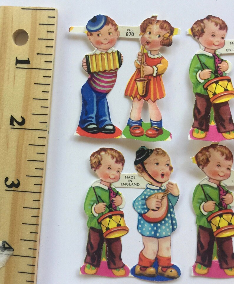 Peut inclure: Six poup&eacute;es en papier vintage repr&eacute;sentant des enfants jouant d'instruments de musique. Les poup&eacute;es sont habill&eacute;es de v&ecirc;tements color&eacute;s et tiennent des instruments tels qu'un tambour, un accord&eacute;on, un saxophone et un banjo. Les poup&eacute;es sont imprim&eacute;es sur du papier blanc et portent une &eacute;tiquette "Made in England".