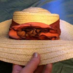 Pu&ograve; includere: Un cappello di paglia a tesa larga con un accento di fiore rosa e bordeaux.