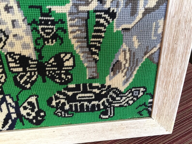 May include: A framed needlepoint piece with a green background and a variety of colorful embroidered designs, including butterflies, insects, and a turtle.