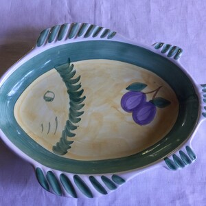 May include: A ceramic fish-shaped serving dish with a green and white border and a yellow interior. The fish has a painted eye and a purple plum with green leaves on the side.